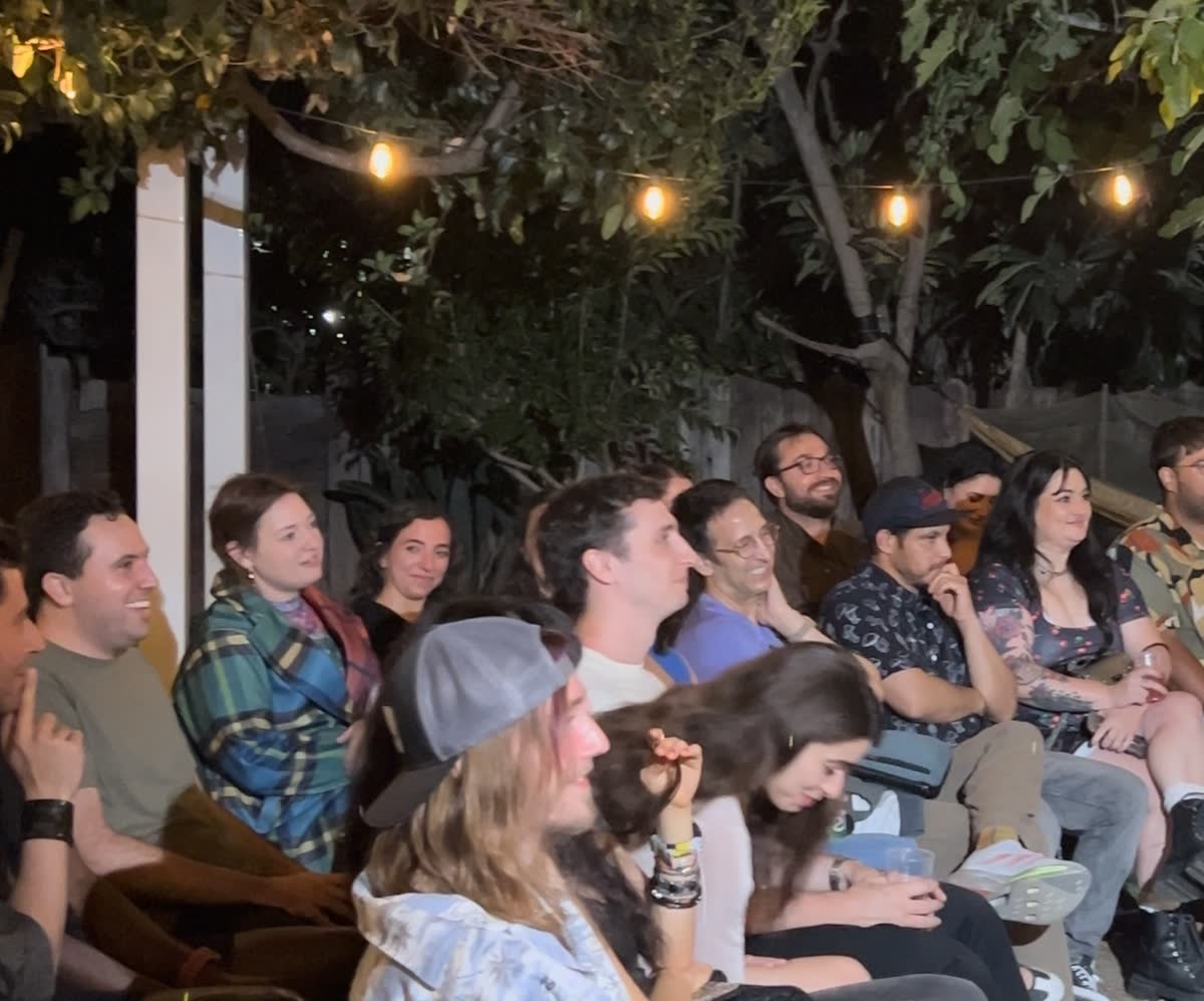 Rows of the audience under the string lights, faces turned the same way and half of them mid-laugh.