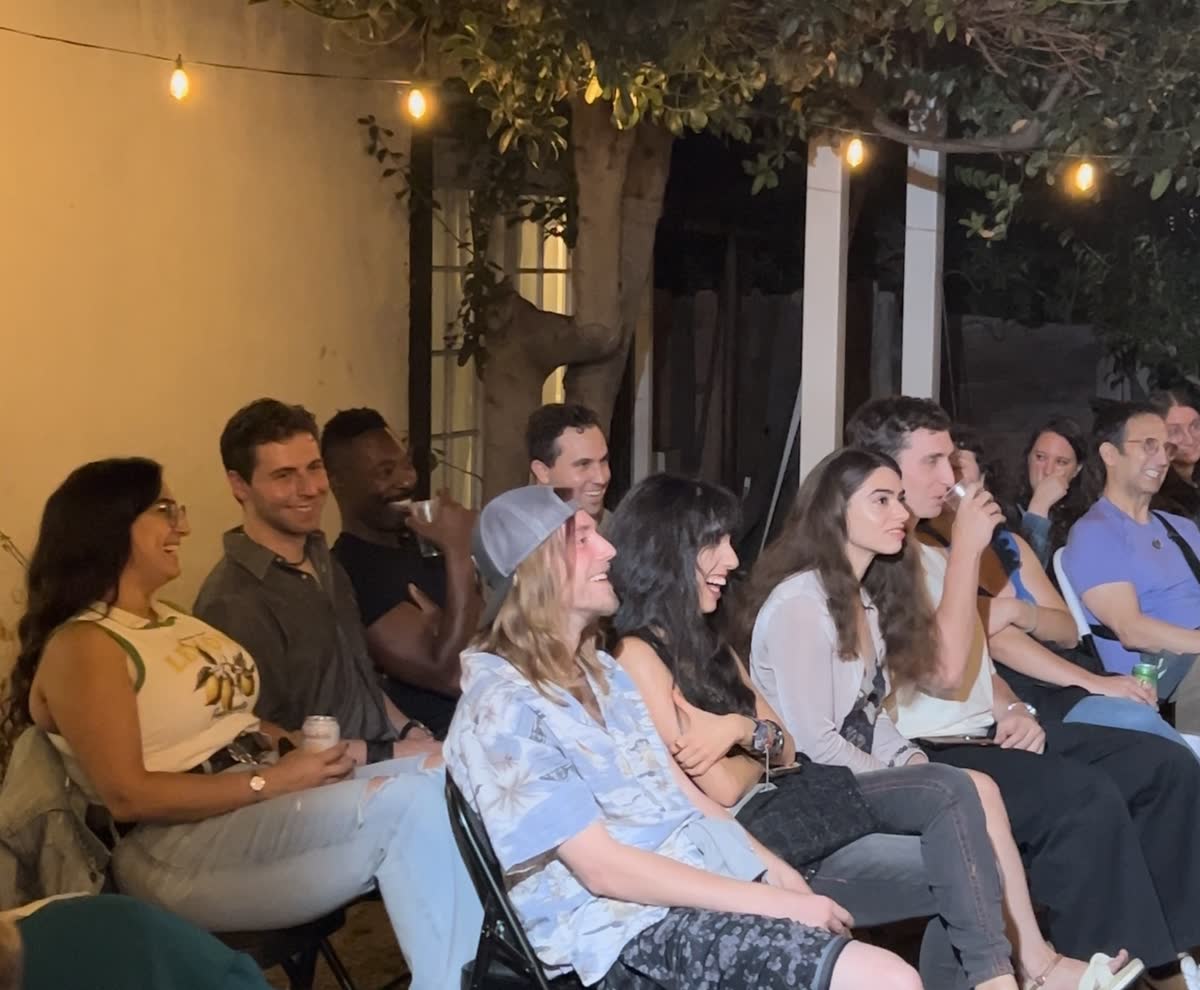 A block of the audience leaning together on folding chairs, drinks in hand, all laughing at once under the bulbs.