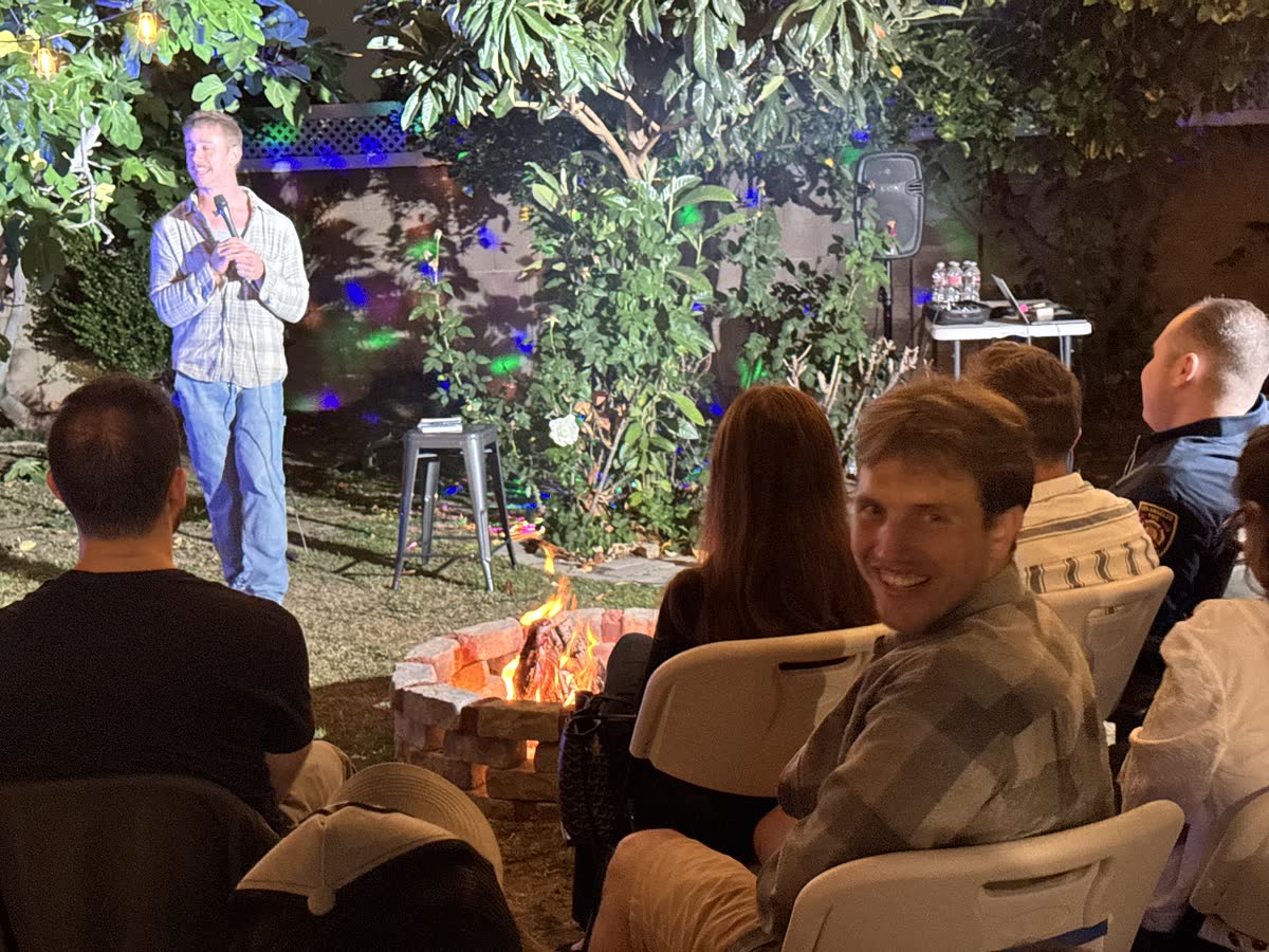 An audience member turning back toward the camera, grinning, while the comic keeps working behind him and the fire burns between the chairs.