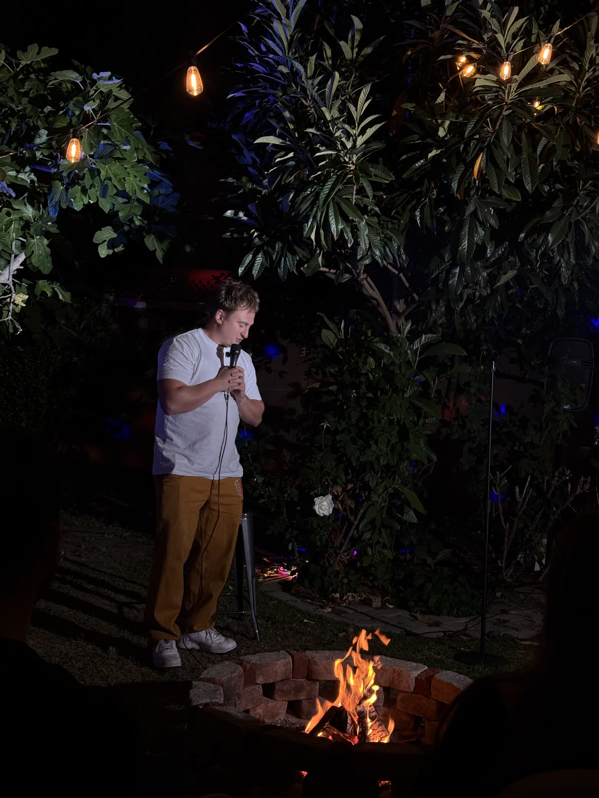 A comic standing quietly on the mic in the dark, head down, the firepit burning at his feet and bare bulbs above him.