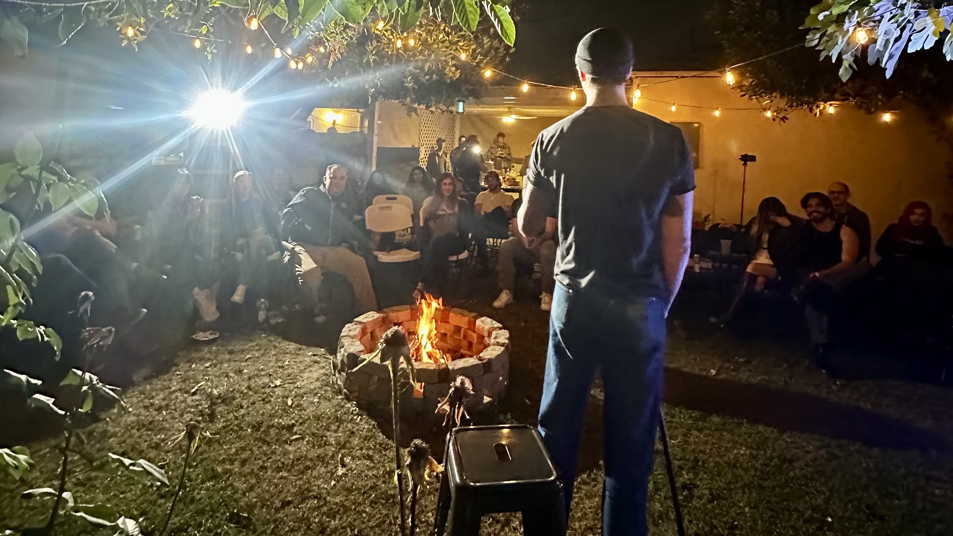 The room seen from behind the comic: a fire burning in the brick pit, the crowd in folding chairs around it, string lights overhead.