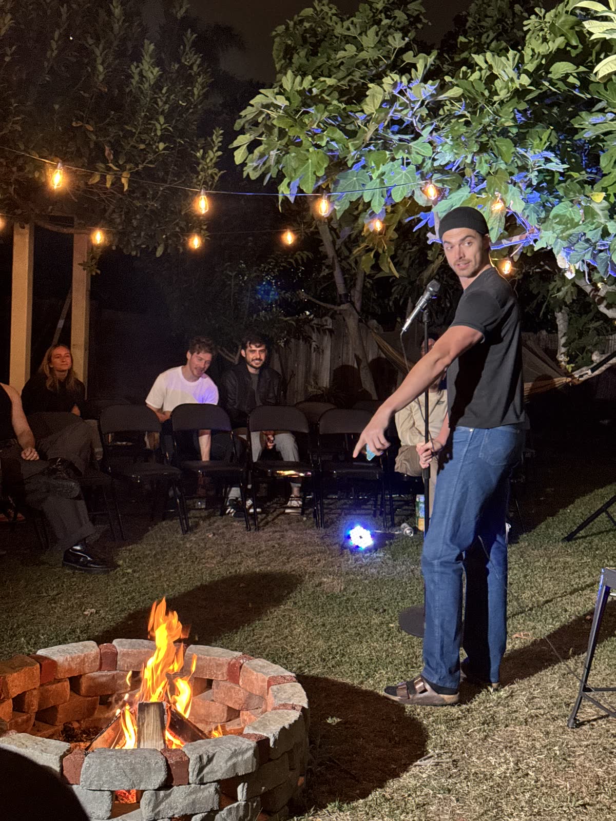A comic in a beanie leaning out of his stance to point at someone off camera, the brick firepit burning in the foreground and empty chairs behind him.