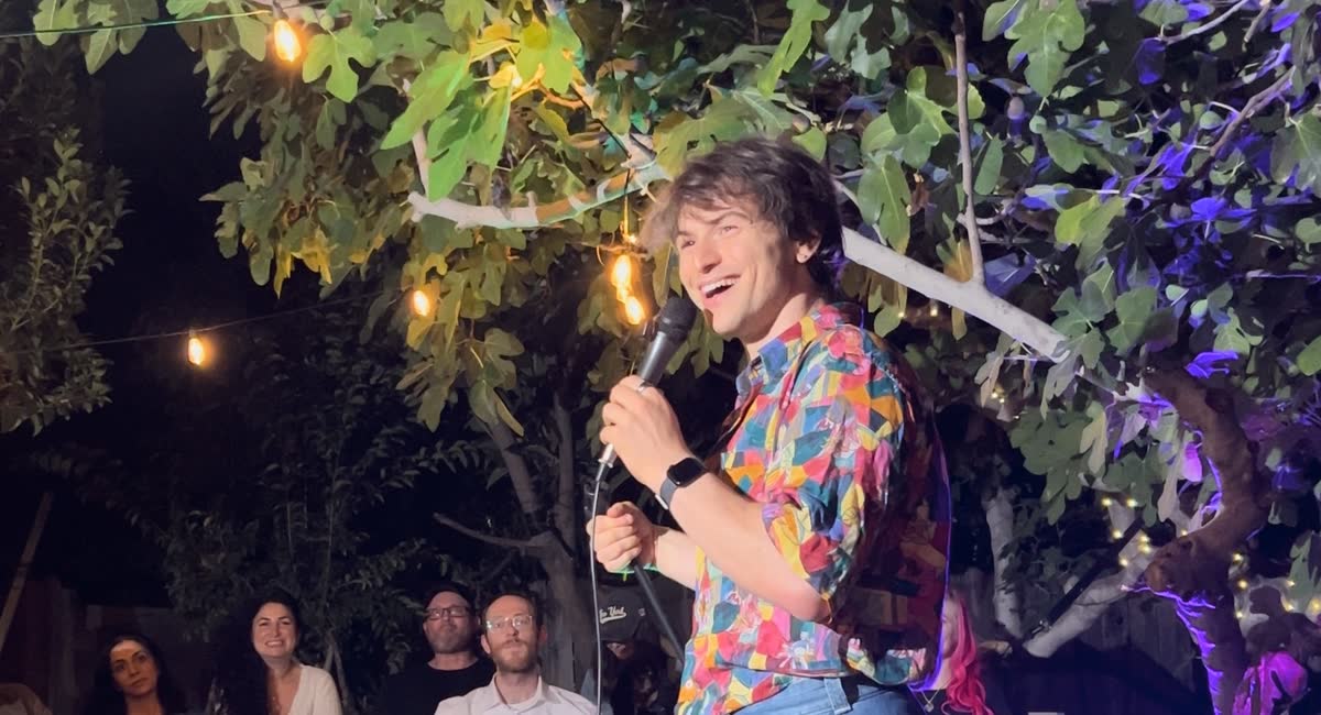 A comic in a bright patterned shirt mid-punchline on the mic, framed by leaves and hanging bulbs, with the front row watching below him.