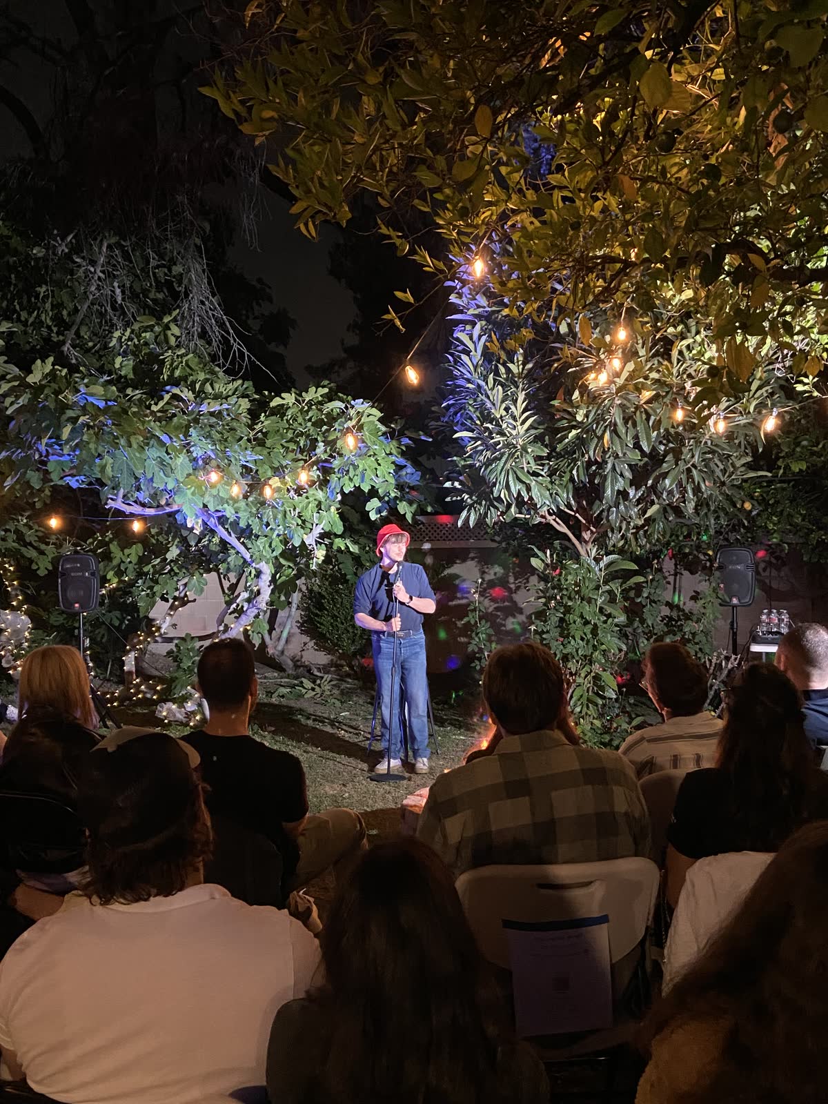 A comic in a red cap on the mic, seen over the heads and shoulders of a packed audience, with lit trees and a speaker behind him.