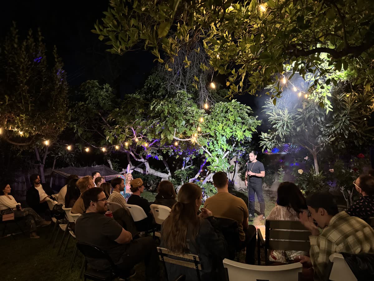 The whole backyard from the back of the room: a full audience on folding chairs under strings of warm bulbs, watching a comic standing on the grass beneath the trees.