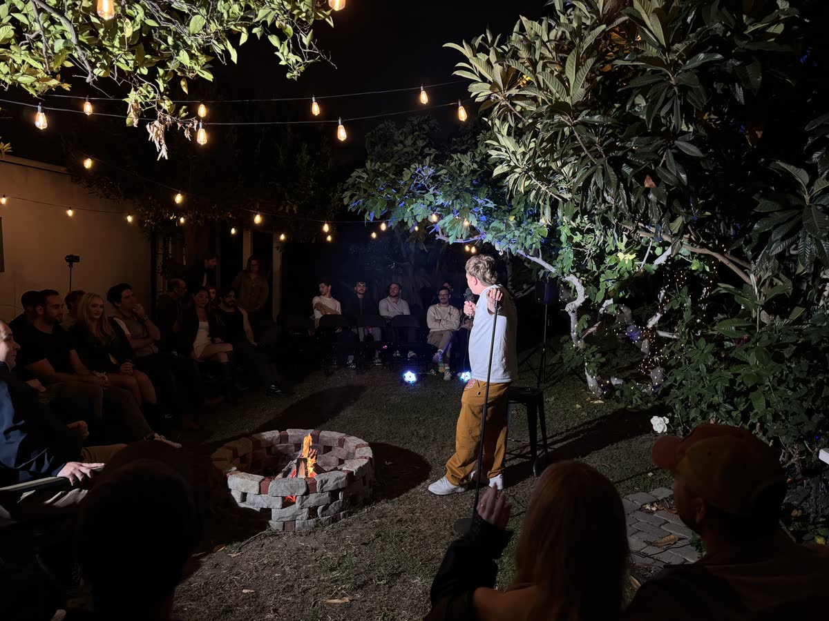 The full room from the side: the ring of chairs, the fire, and a comic on the mic under a canopy of string lights.